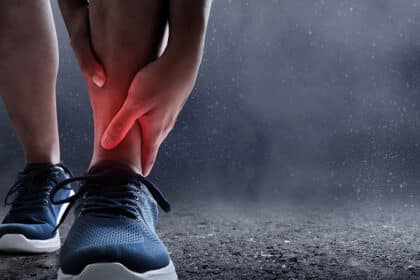 Person hält sich den Knöchel vor Schmerz, rote Markierung deutet auf eine Verletzung oder Verstauchung hin.