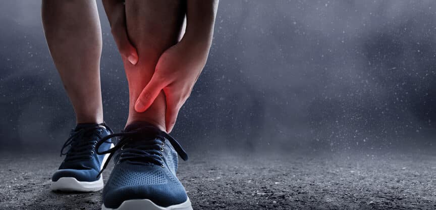 Person hält sich den Knöchel vor Schmerz, rote Markierung deutet auf eine Verletzung oder Verstauchung hin.