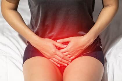 Frau sitzt auf dem Bett und hält sich mit beiden Händen den Unterbauch, rotes Licht signalisiert Schmerz.