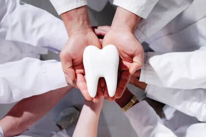 Mehrere Hände in weißen Kitteln halten gemeinsam ein großes Zahnmodell als Symbol für Teamarbeit.