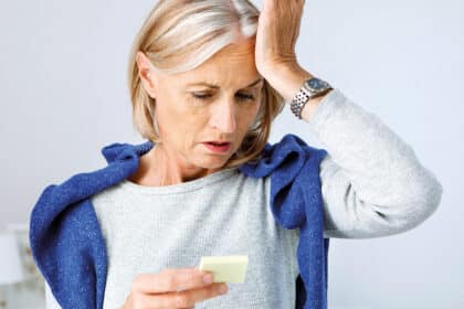 Ältere Frau hält sich die Stirn und schaut besorgt auf einen Notizzettel, wirkt nachdenklich oder vergesslich.