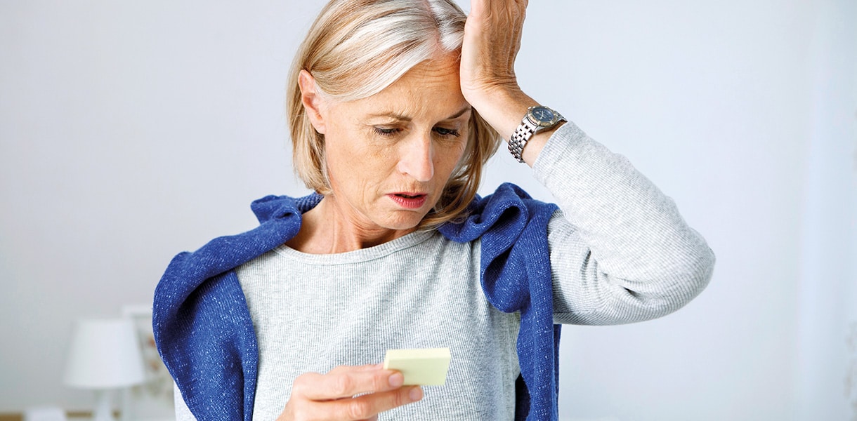 Ältere Frau hält sich die Stirn und schaut besorgt auf einen Notizzettel, wirkt nachdenklich oder vergesslich.