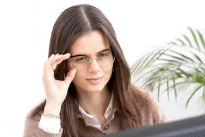 Junge Frau mit Brille blickt konzentriert auf einen Bildschirm, helle Umgebung mit Pflanze im Hintergrund.