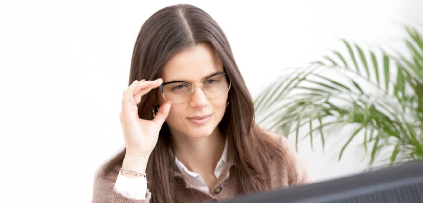 Junge Frau mit Brille blickt konzentriert auf einen Bildschirm, helle Umgebung mit Pflanze im Hintergrund.