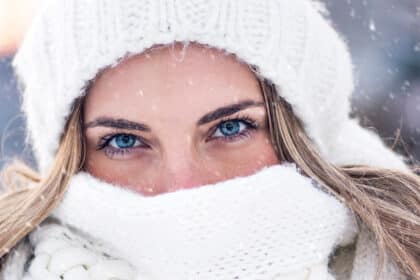 Nahaufnahme einer Frau im Winter, eingehüllt in weiße Mütze und Schal, mit Schneeflocken im Hintergrund.