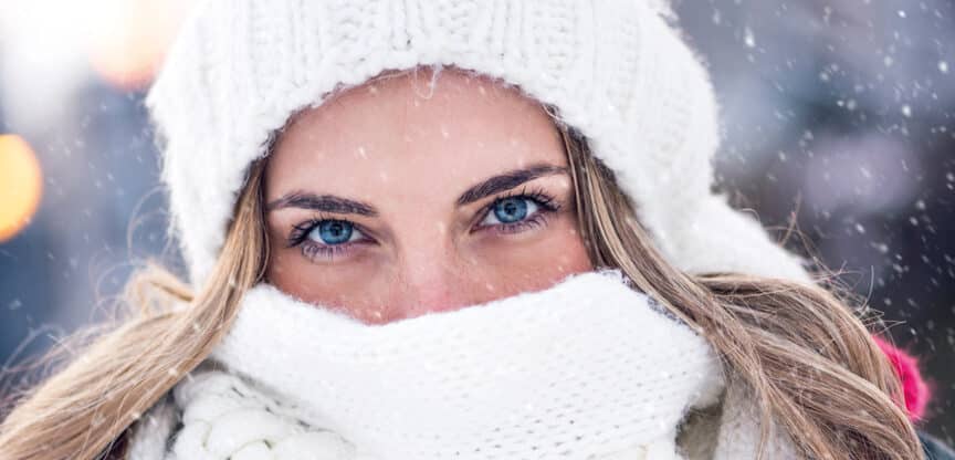 Nahaufnahme einer Frau im Winter, eingehüllt in weiße Mütze und Schal, mit Schneeflocken im Hintergrund.