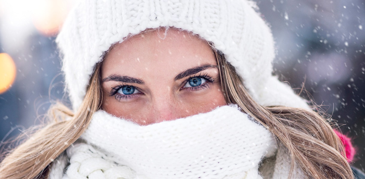 Nahaufnahme einer Frau im Winter, eingehüllt in weiße Mütze und Schal, mit Schneeflocken im Hintergrund.