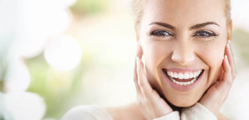 Nahaufnahme einer lächelnden Frau mit gesunden Zähnen, die ihr Gesicht mit beiden Händen berührt.