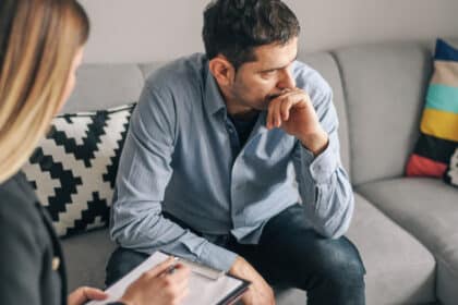 Mann sitzt nachdenklich auf dem Sofa und spricht mit einer Therapeutin, die sich Notizen macht.