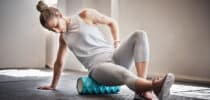 Frau sitzt auf einer Faszienrolle und massiert ihren unteren Rücken, wirkt angespannt oder schmerzgeplagt.