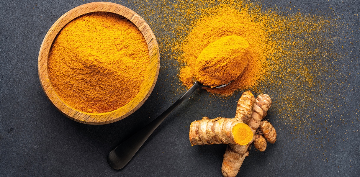 Schale und Löffel mit Kurkumapulver sowie frische Kurkumawurzeln auf dunklem Hintergrund.