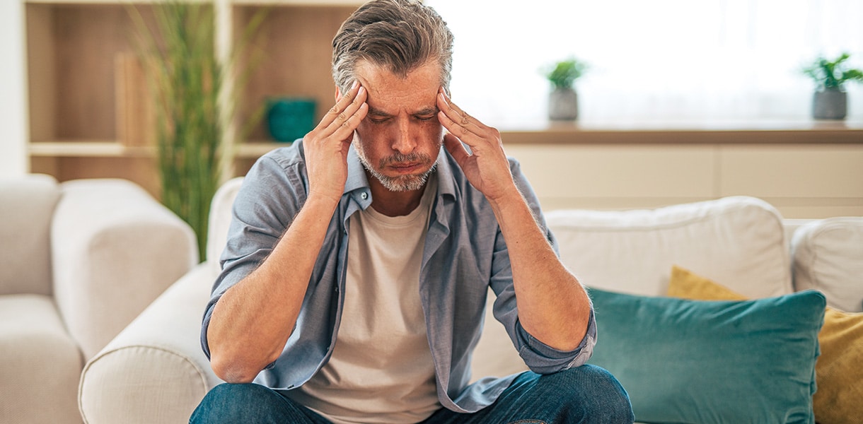 Mann mittleren Alters sitzt auf einem Sofa und hält sich mit beiden Händen die Schläfen.
