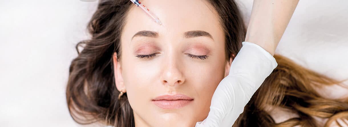 Frau liegt mit geschlossenen Augen, während eine kosmetische Injektion an der Stirn vorbereitet wird.