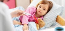Krankes Kind liegt mit Kuscheltier im Bett, während die Körpertemperatur gemessen wird.