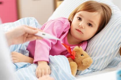 Krankes Kind liegt mit Kuscheltier im Bett, während die Körpertemperatur gemessen wird.