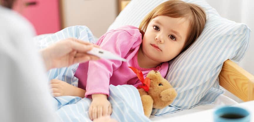 Krankes Kind liegt mit Kuscheltier im Bett, während die Körpertemperatur gemessen wird.