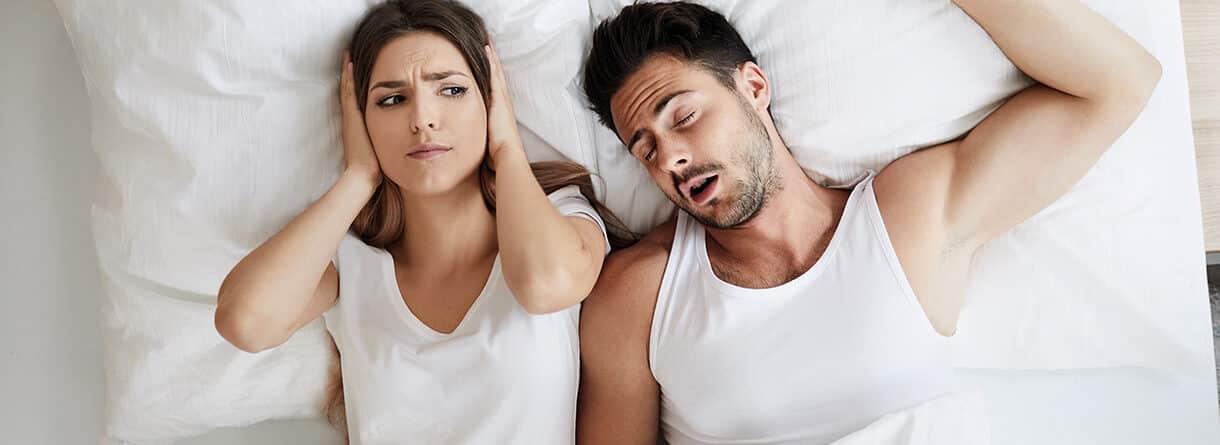 Frau hält sich im Bett die Ohren zu, während ihr Partner neben ihr laut schnarcht.