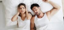 Frau hält sich im Bett die Ohren zu, während ihr Partner neben ihr laut schnarcht.