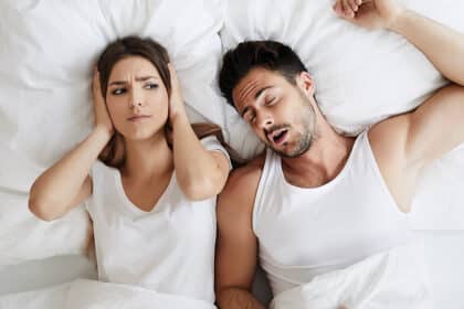 Frau hält sich im Bett die Ohren zu, während ihr Partner neben ihr laut schnarcht.
