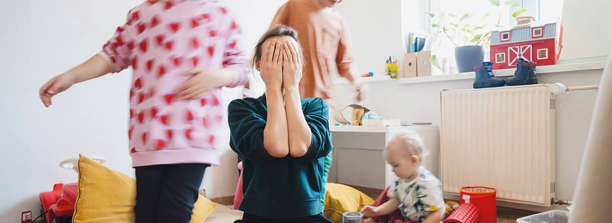 Überforderte Mutter sitzt zwischen spielenden Kindern und hält sich erschöpft die Hände vors Gesicht.