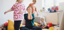 Überforderte Mutter sitzt zwischen spielenden Kindern und hält sich erschöpft die Hände vors Gesicht.