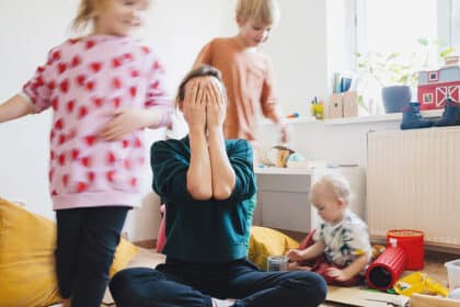 Überforderte Mutter sitzt zwischen spielenden Kindern und hält sich erschöpft die Hände vors Gesicht.