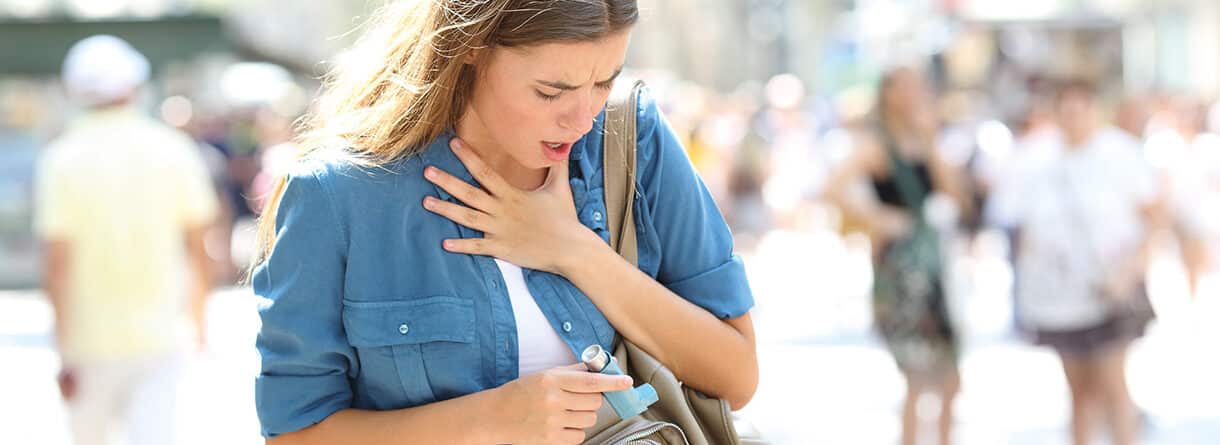 Junge Frau mit Atembeschwerden hält sich an den Hals und trägt einen Asthma-Inhalator in der Hand.