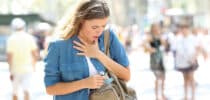 Junge Frau mit Atembeschwerden hält sich an den Hals und trägt einen Asthma-Inhalator in der Hand.