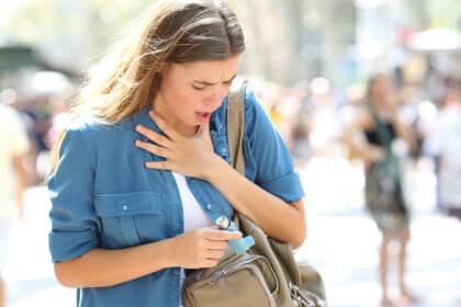Junge Frau mit Atembeschwerden hält sich an den Hals und trägt einen Asthma-Inhalator in der Hand.