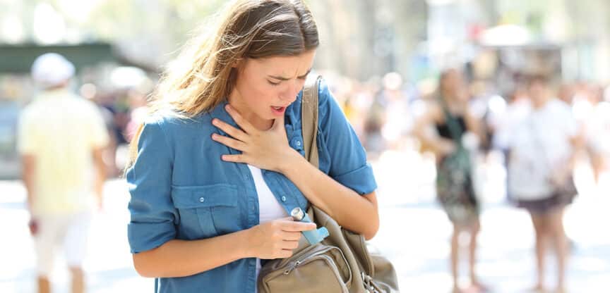 Junge Frau mit Atembeschwerden hält sich an den Hals und trägt einen Asthma-Inhalator in der Hand.