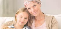 Krebskranke Mutter mit Kopftuch sitzt eng umschlungen mit ihrer Tochter auf dem Sofa.