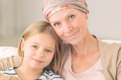 Krebskranke Mutter mit Kopftuch sitzt eng umschlungen mit ihrer Tochter auf dem Sofa.