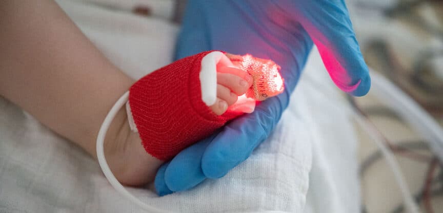 Nahaufnahme eines Neugeborenen mit verbundenem Fuß und leuchtendem Sensor, gehalten von einer behandschuhten Hand.