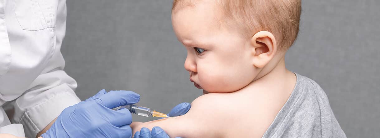 Nahaufnahme eines Babys, das von medizinischem Fachpersonal mit einer Spritze in den Oberarm geimpft wird.