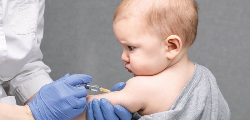 Nahaufnahme eines Babys, das von medizinischem Fachpersonal mit einer Spritze in den Oberarm geimpft wird.